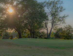 The sun sets behind large trees on the grassy fairways of Spring Lake Country Club, casting long shadows over the green as the sky glows softly in the evening light.