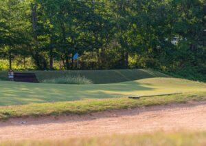 A golf green with a blue flag on the hole at Spring Lake Country Club, bordered by sand and lush trees in the background on a sunny day.