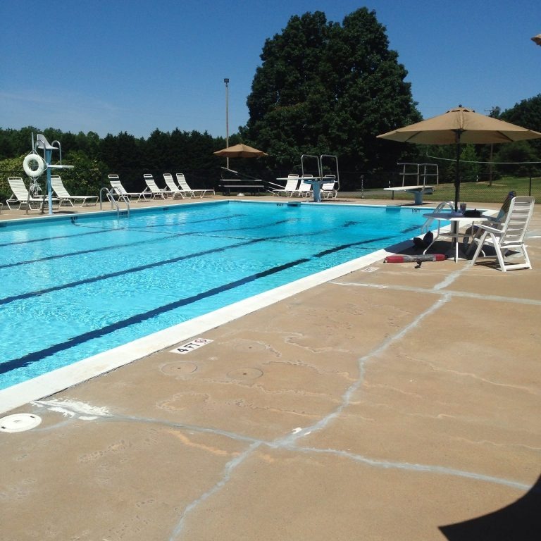 An outdoor swimming pool with lane markings at Spring Lake Country Club, surrounded by lounge chairs and umbrellas. Lifeguard chairs and trees are visible in the background under a clear blue sky.