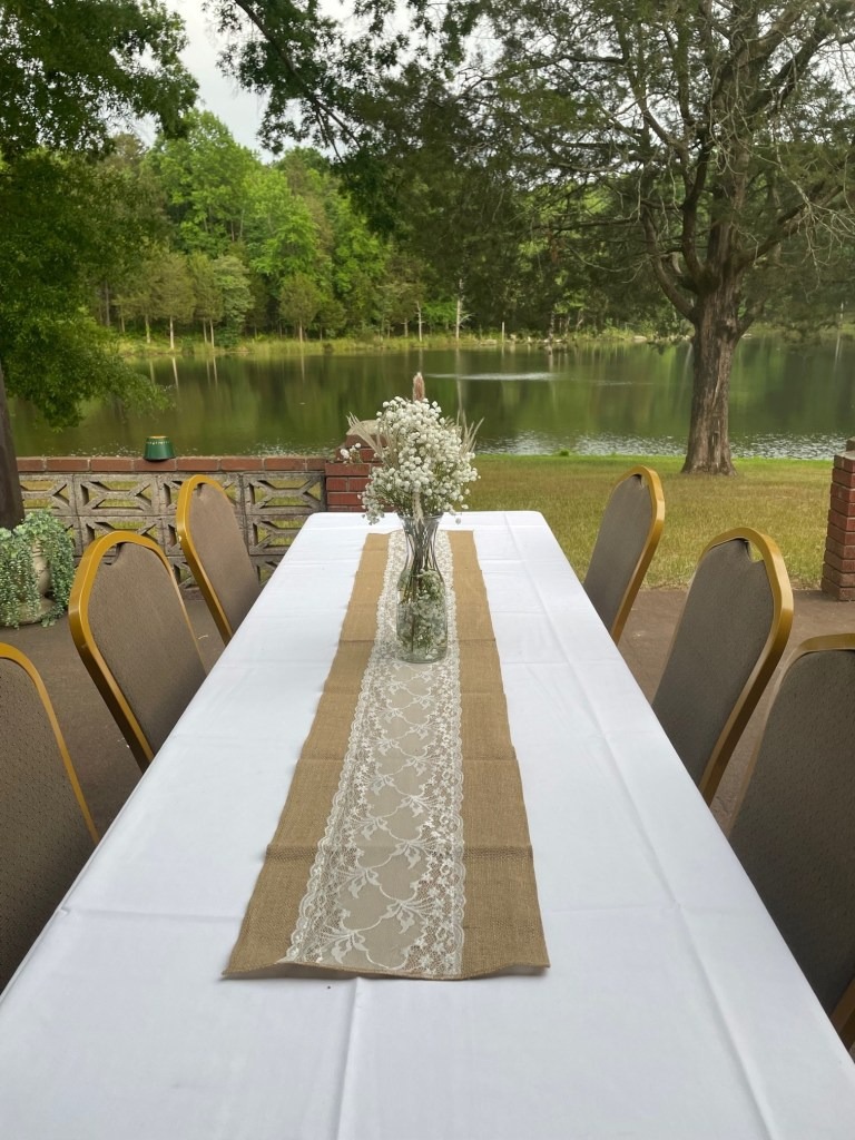 A long table with a white tablecloth and lace runner is set outdoors by the lake at Spring Lake Country Club, surrounded by chairs. A vase with white flowers sits as a centerpiece, with trees and water visible in the background.