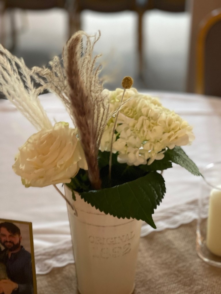 A white tin container with “ORIGINAL 1962”—filled with white roses, hydrangeas, greenery, and dried grasses—sits on a table at Spring Lake Country Club, beside a candle and a blurred photo in the background.