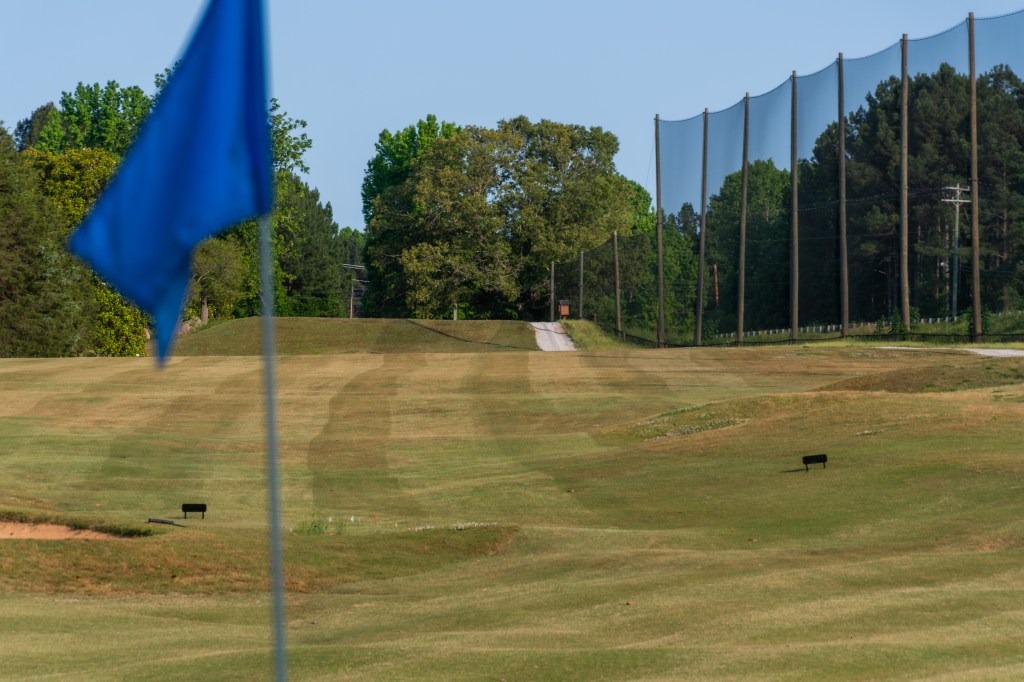 A blue flag in the foreground marks a section of Spring Lake Country Club’s golf course, with green fairways, a tree-lined edge, and a tall netting fence on the right under a clear sky.