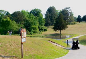A golfer swings a club near a pond at Spring Lake Country Club, while another person sits in a golf cart on a paved path. The scene is surrounded by trees and grassy hills under a clear sky.
