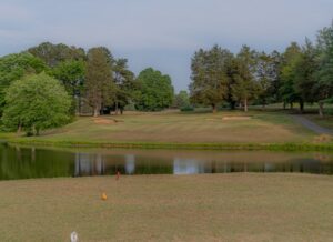 A grassy golf course at Spring Lake Country Club features sand bunkers, is bordered by trees, and has a small pond in the foreground under a pale blue sky.