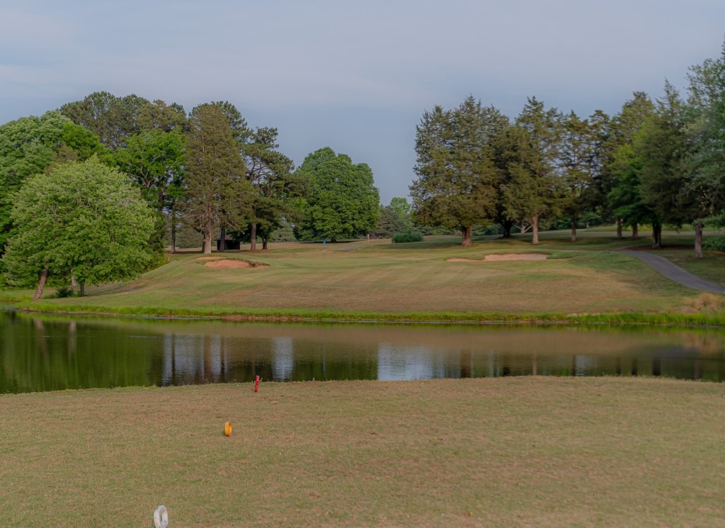 A grassy golf course at Spring Lake Country Club features sand bunkers, is bordered by trees, and has a small pond in the foreground under a pale blue sky.