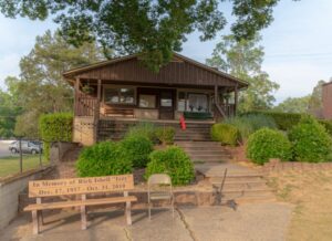 A brown wooden house with stairs leading up to a porch at Spring Lake Country Club, surrounded by green bushes. In front is a bench with a memorial plaque for Rick Isbell Izzy (1957–2010), and a single chair beside it.