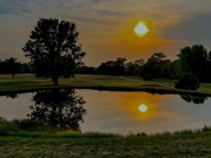 A serene sunset scene at Spring Lake Country Club with a glowing sun in a cloudy sky, reflected in a calm pond. Tall trees cast silhouettes and reflections on the water, while grass borders the pond in the foreground.