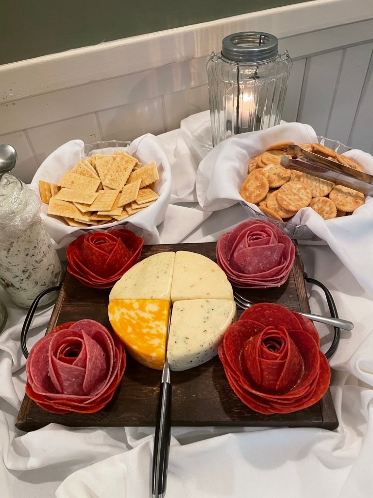 A platter with sliced cheese and salami arranged as roses, surrounded by serving knives. Behind are bowls of crackers, a jar of herb cheese spread, and a lit candle—all set for an elegant Spring Lake Country Club gathering.
