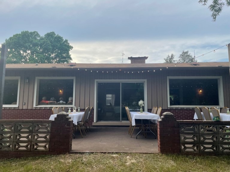 A backyard patio at Spring Lake Country Club with tables and chairs covered in white tablecloths, set for an outdoor gathering in front of a one-story house with string lights above and large windows.