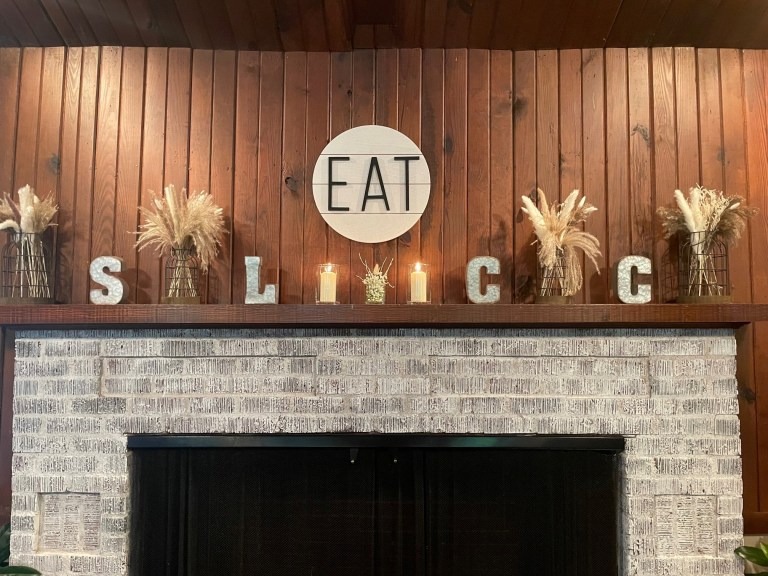 A rustic mantel at Spring Lake Country Club features a whitewashed brick fireplace, wooden backdrop, and decor spelling SLICE. Above are vases with dried wheat, three lit candles, and a round sign with EAT in bold black letters.