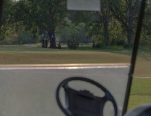 View from inside a golf cart at Spring Lake Country Club, with the steering wheel in the foreground and a lush green golf course, trees, and a distant golfer visible through the windshield.