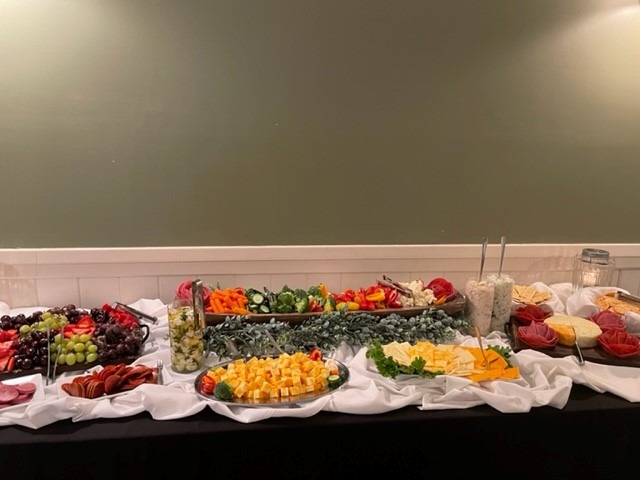 A buffet table at Spring Lake Country Club features assorted cheeses, sliced meats, grapes, crackers, fresh vegetables, and dips arranged on trays over a white tablecloth against a green wall.