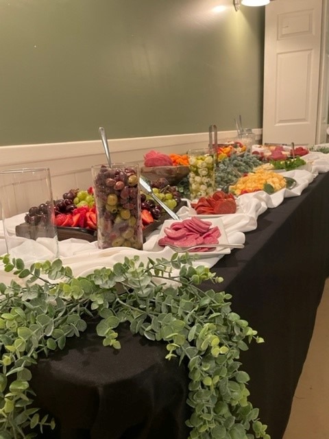 A long buffet table at Spring Lake Country Club is covered with a black tablecloth and white cloth accents, displaying assorted fruits, cheeses, and meats, with serving utensils and green leafy garland as decoration.