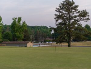 A golf green with a white flag in the foreground sits beneath cloudy skies at Spring Lake Country Club, with large trees, a small building, and a fenced swimming pool visible in the background.