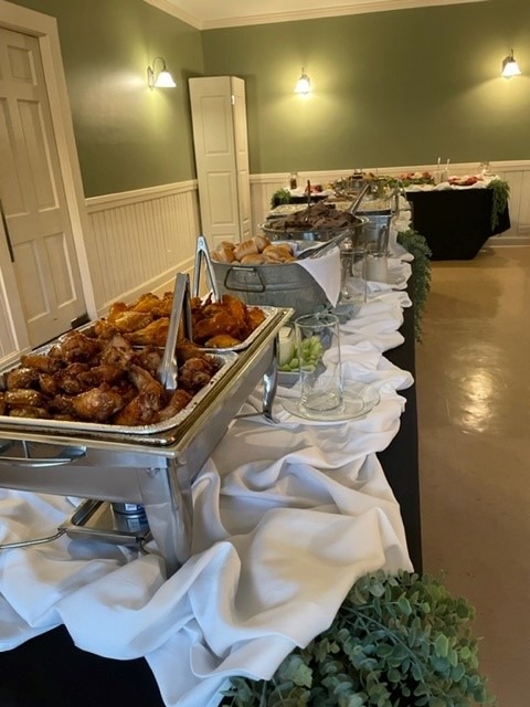 A buffet table at Spring Lake Country Club features trays of chicken wings, rolls, and assorted dishes on white tablecloths with greenery accents, set in a room with green walls and soft lighting.