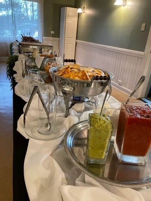 A buffet table at Spring Lake Country Club is set with covered chafing dishes, serving utensils, and trays of condiments, including green relish and red sauce, in a softly lit room with white wainscoting and large windows.