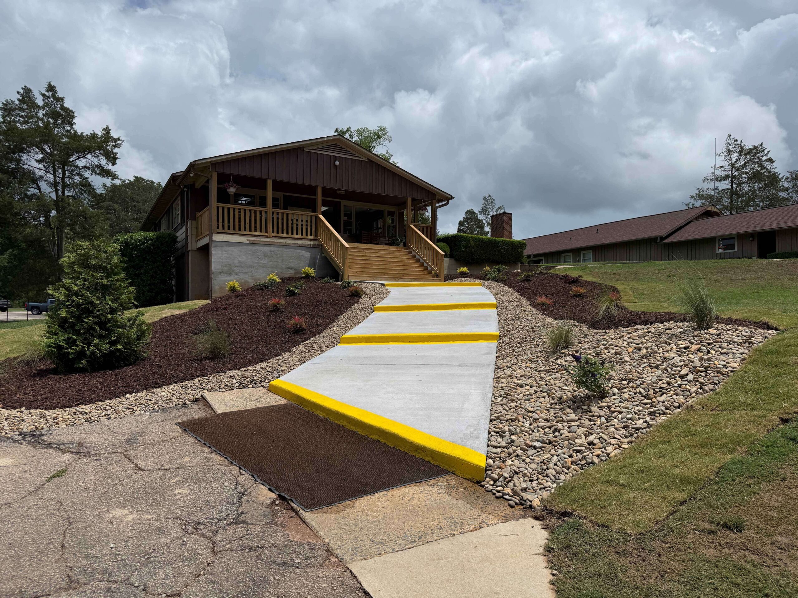 A concrete wheelchair ramp with bright yellow edges leads up to a wooden building at Spring Lake Country Club. The ramp is bordered by rocks, mulch, and small landscaped plants beneath a cloudy, overcast sky.