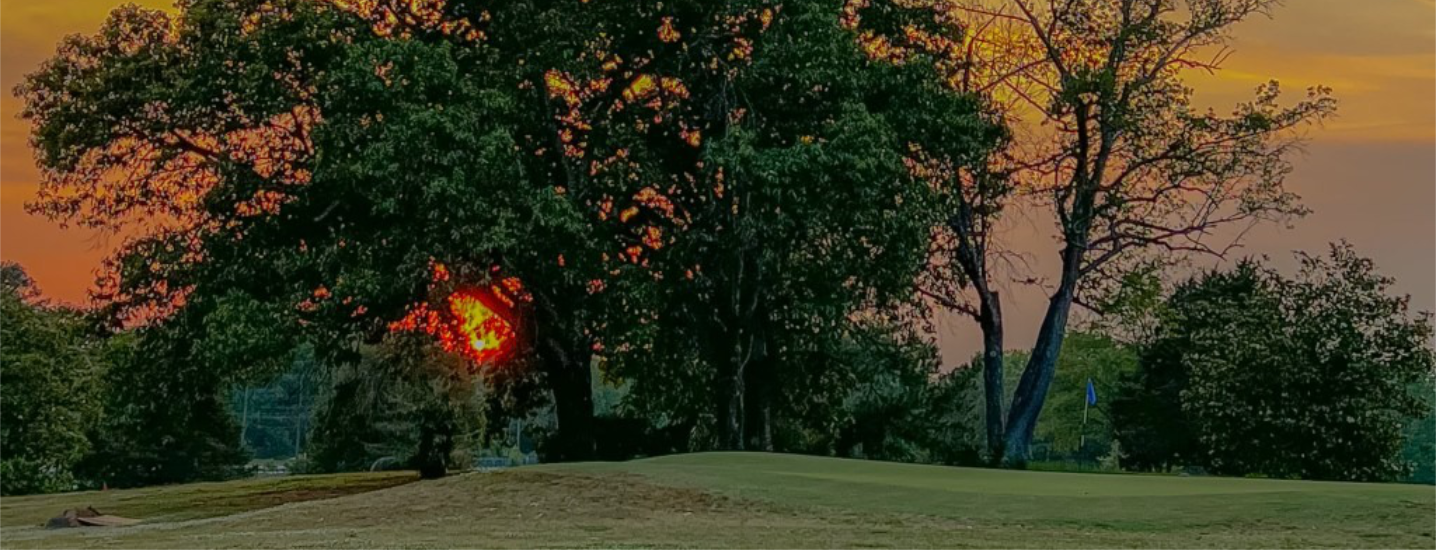 The sun sets behind large trees at Spring Lake Country Club, casting an orange and pink glow across the sky, with green grass and a sand trap visible in the foreground.