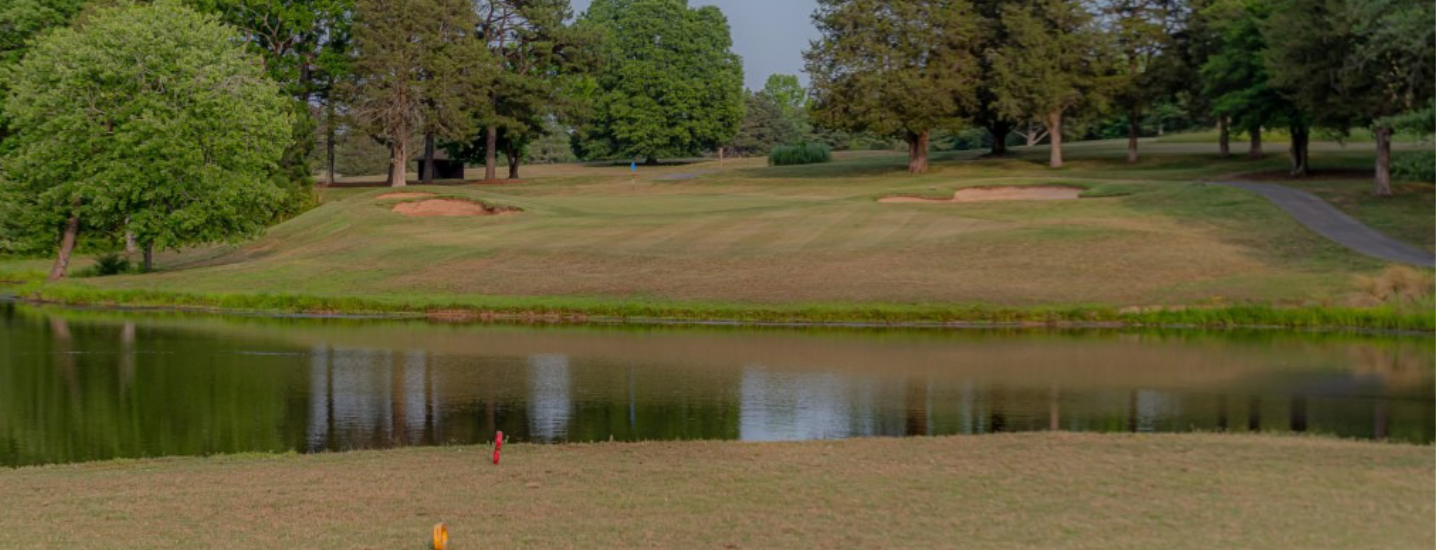 A view across a pond toward the Spring Lake Country Club golf course green with sand bunkers, surrounded by trees and grass under a partially cloudy sky.